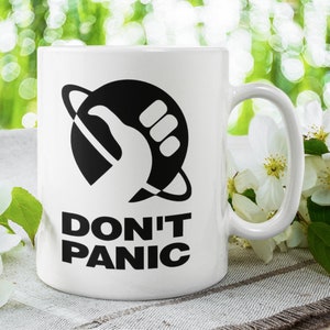 May include: White ceramic mug featuring a black and white graphic of a hand giving a thumbs up inside a circle with a ring around it. The text "DON'T PANIC" is printed below the graphic.