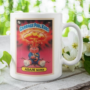 Puede incluir: Taza de cerámica blanca con un diseño de tarjeta de intercambio de Garbage Pail Kids colorido con un personaje de dibujos animados llamado Adam Bomb. La tarjeta tiene un fondo rojo con una explosión amarilla y naranja y el texto "Garbage Pail Kids" y "Adam Bomb".