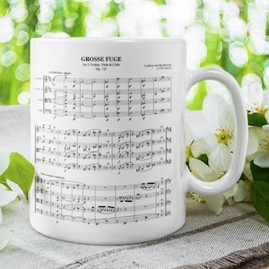 May include: White ceramic coffee mug with a black and white image of sheet music for "Grosse Fuge" by Ludwig van Beethoven. The mug is sitting on a white and brown surface with white flowers.