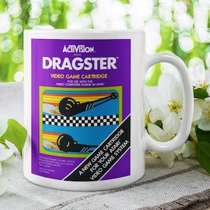 May include: White ceramic mug featuring the "Dragster" video game cartridge design. The mug has a purple background with the game's logo and graphics. The mug also has the text "A NEW GAME CARTRIDGE FOR YOUR ATARI VIDEO GAME SYSTEM".