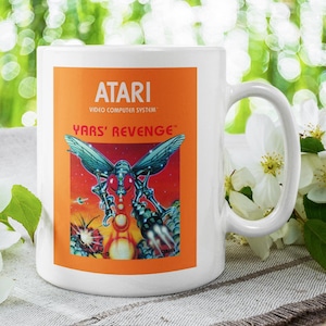 May include: White ceramic mug featuring the Atari Video Computer System logo and the game title "Yars' Revenge" on an orange background. The mug's design includes a colorful graphic of a creature with wings. The mug has a white handle.