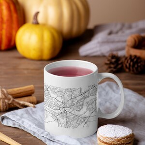 May include: White ceramic mug with a black and white city map design. The mug is filled with pink tea and is sitting on a gray linen napkin with a sugar cookie beside it.