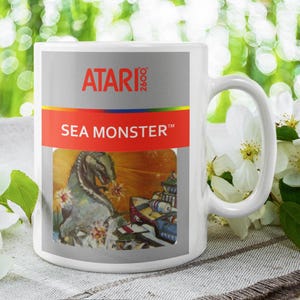 May include: White ceramic mug featuring the Atari 2600 logo and the text "SEA MONSTER". The mug displays a retro video game graphic of a sea monster attacking a ship. The mug is set against a blurred floral background.