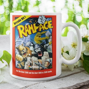 May include: White ceramic mug featuring the Rampage arcade game artwork. The design includes the game's title, a monster-themed illustration, and the text "Arcade Hit!". The mug is a classic coffee cup shape.
