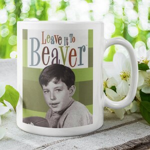 May include: White ceramic mug featuring a vintage image of a young boy and the text "Leave it to Beaver" in colorful letters. The mug has a curved handle and is set against a blurred green background with white flowers.