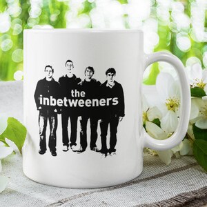 May include: White ceramic mug with a black and white image of the band "The Inbetweeners" printed on the front. The band members are standing in a row, and the band name is written in bold letters below them.
