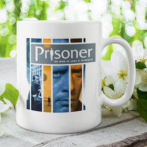 May include: White ceramic mug featuring a design from "The Prisoner" TV series. The design includes the show's title and the phrase "No man is just a number" with images of the main character. The mug has a curved handle.
