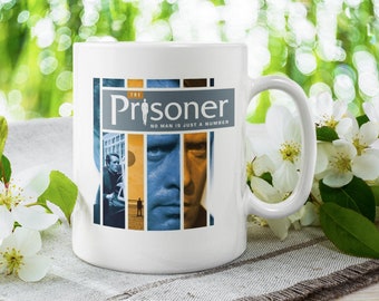 May include: White ceramic mug featuring a design from "The Prisoner" TV series. The design includes the show's title and the phrase "No man is just a number" with images of the main character. The mug has a curved handle.