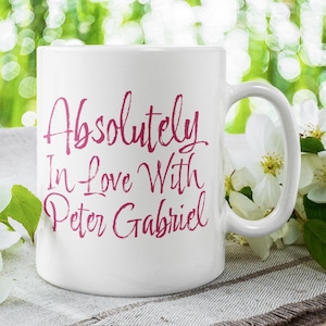 Può includere: Tazza in ceramica bianca con la scritta "Absolutely In Love With Peter Gabriel" in rosa. La tazza è presentata con fiori bianchi e foglie verdi sullo sfondo.