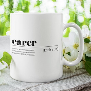Könnte beinhalten: Eine weiße Keramiktasse mit einer schwarzen Grafik, auf der "carer" und seine Definition steht. Die Tasse steht auf einer weißen und braunen gestreiften Oberfläche mit weißen Blumen im Hintergrund.