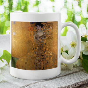 Może przedstawiać: Biały ceramiczny kubek z fragmentem obrazu Gustava Klimta, "Portret Adele Bloch-Bauer I". Dzieło przedstawia kobietę w bogato zdobionej złotej sukni na złotym tle. Kubek ma zakrzywiony uchwyt i jest otoczony białymi kwiatami.
