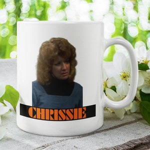 May include: White ceramic mug with a picture of a woman with blonde hair and the word "CHRISSIE" printed in orange letters on a black background.