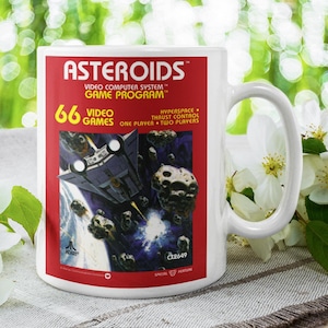 Puede incluir: Taza de cerámica blanca con el videojuego Asteroids. La taza tiene una etiqueta roja con el logotipo del juego y un gráfico de una nave espacial y asteroides. La taza también tiene el texto "66 Video Games".