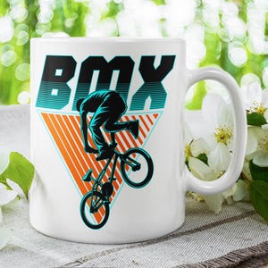 以下が含まれることがあります： 白地に黒と青のBMXに乗る人のグラフィックが描かれた白いセラミックマグカップ。グラフィックは三角形で、その上に「BMX」の文字が書かれている。