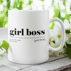 Może przedstawiać: Biały ceramiczny kubek z czarnym tekstem, który brzmi "girl boss" i definicją tego terminu. Kubek stoi na białej i brązowej, teksturowanej powierzchni z białymi kwiatami.