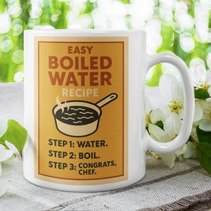 May include: White ceramic mug featuring a humorous "Easy Boiled Water Recipe". The recipe is printed on a tan background and includes three steps. The mug is surrounded by white flowers and green foliage.