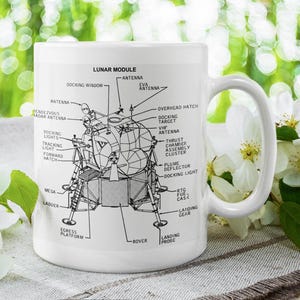 May include: White ceramic mug featuring a detailed diagram of the Lunar Module, with labels like "Docking Window" and "Landing Gear." The mug is set against a backdrop of white flowers and green foliage.