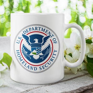 May include: White ceramic mug with a blue and red seal of the U.S. Department of Homeland Security. The seal features a bald eagle with wings spread and a shield on its chest. The mug is sitting on a white and gray textured surface.