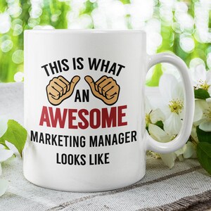 Może przedstawiać: Biały ceramiczny kubek do kawy z czarno-czerwonym wzorem, na którym widnieje napis "Tak wygląda niesamowity menedżer marketingu". Kubek ma po obu stronach słowa "niesamowity" dwa uniesione kciuki.