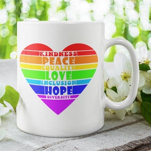 Op de afbeelding: Witte keramische mok met een regenboogvormig hartontwerp. Het hart bevat de woorden "kindness", "peace", "equality", "love", "inclusion", "hope" en "diversity".
