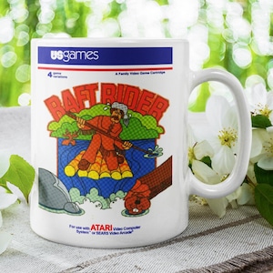 May include: White ceramic mug featuring the Raft Rider video game design. The mug displays a colorful illustration of a man on a raft, with the game title and "us games" logo. The mug is a standard size with a curved handle.