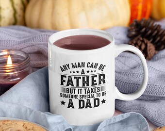 May include: White ceramic mug with a black graphic that reads "Any man can be a father, but it takes someone special to be a dad." The mug is filled with a brown liquid and is sitting on a blue knitted blanket with a cinnamon stick and a pine cone.