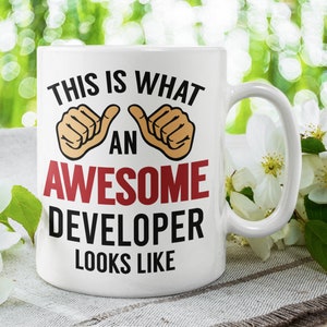 May include: White ceramic coffee mug with a black and red design that says "This is what an awesome developer looks like". The mug has two thumbs up on either side of the text. The mug is sitting on a white surface with white flowers.