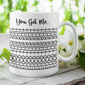 Może przedstawiać: Biały ceramiczny kubek z napisem "You Get Me..." czarną czcionką. Kubek ma czarny geometryczny wzór na korpusie. Kubek jest ustawiony na rozmytym zielonym tle z białymi kwiatami.