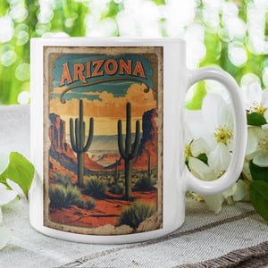 May include: White ceramic mug with a vintage Arizona landscape design. The design features the word "Arizona" in an arching font, with cacti and a desert scene. The mug has a curved handle and is suitable for hot drinks.