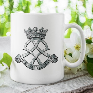 May include: White ceramic mug displaying The Royal Yeomanry emblem. The design includes a crown, intertwined elements, and a banner with text. The mug is presented with white flowers and green foliage in the background.