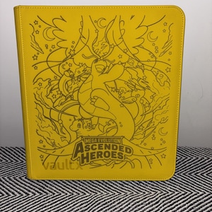 May include: A bright yellow rectangular trading card binder featuring a dark line art illustration of various creatures and celestial motifs. The text "MEGA EVOLUTION ASCENDED HEROES" is printed in the center, with the brand name "vaultx" visible below.