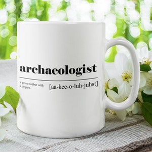 Op de afbeelding: Een witte keramische mok met een zwarte inscriptie die "archeoloog" luidt en een definitie van het woord. De mok staat op een wit en bruin stoffen oppervlak met witte bloemen.