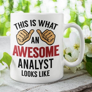 Op de afbeelding: Een witte keramische koffiemok met een zwart en rood ontwerp met de tekst "Dit is hoe een geweldige analist eruitziet". De mok heeft aan beide kanten van de tekst twee duimen omhoog symbolen. De mok staat op een wit oppervlak met witte bloemen.