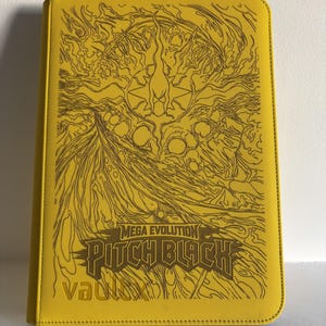 May include: A bright yellow rectangular card binder featuring a dark brown line art illustration of a creature and swirling energy patterns. The text reads Mega Evolution, Pitch Black, and Vaultx in a stylized font on the front cover.