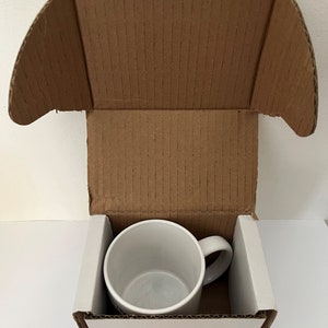 May include: A white ceramic mug with a handle is inside a cardboard box with a white interior. The box is open and the mug is visible.