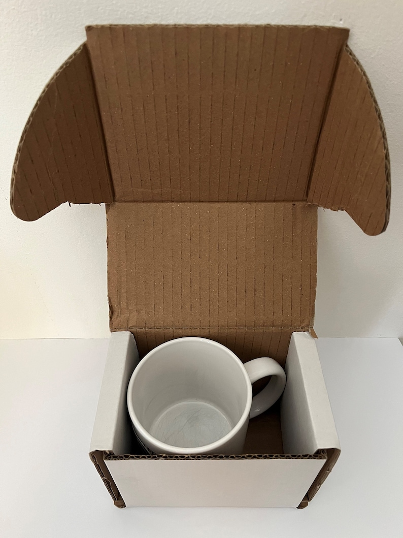 May include: A white ceramic mug with a handle is inside a cardboard box with a white interior. The box is open and the mug is visible.