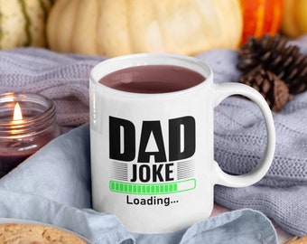 May include: White ceramic mug with a black and green design that says "Dad Joke Loading..." with a progress bar. The mug is filled with a brown liquid and is sitting on a blue knitted blanket.