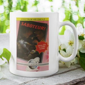 Puede incluir: Taza de cerámica blanca con un diseño de videojuego retro. La taza muestra el título del juego "Saboteur!" con un gráfico vintage de una escena de artes marciales. La taza tiene un asa curva y está sobre un fondo floral.