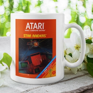 May include: White ceramic mug featuring the Atari Video Computer System logo and the text "Star Raiders" on an orange background. The mug has a wraparound design with a space-themed graphic. The mug is set against a backdrop of flowers.