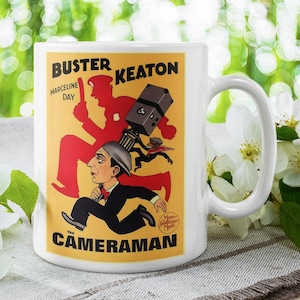 Op de afbeelding: Witte keramische mok met een vintage filmposterontwerp voor "The Cameraman" met Buster Keaton. De posterkunst bevat een rennende figuur, een camera en de namen "Buster Keaton" en "Marceline Day".