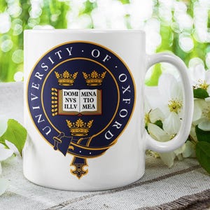 May include: White ceramic mug displaying the University of Oxford emblem in navy and gold. The emblem is circular, with text and a central crest. The mug has a curved handle. White flowers are arranged around the mug.