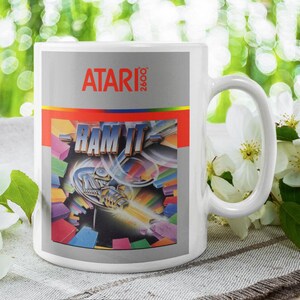 May include: White ceramic mug featuring the Atari 2600 logo and the "RAM IT" game art. The mug has a large handle and is set against a blurred background with white flowers and greenery.