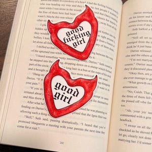 May include: Two heart-shaped stickers with a red outline and white center, featuring the words "good fucking girl" and "good girl" in a bold, black font. The stickers are placed on the open pages of a book.