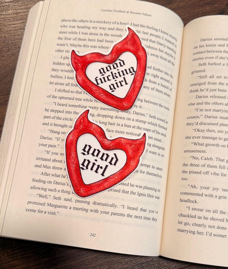 May include: Two heart-shaped stickers with a red outline and white center, featuring the words "good fucking girl" and "good girl" in a bold, black font. The stickers are placed on the open pages of a book.