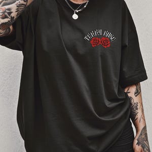 Peut inclure: T-shirt noir surdimensionn&eacute; avec le texte "Terra Rose" au-dessus de deux illustrations de roses rouges. Le t-shirt a des manches courtes et une coupe ample. Le motif est centr&eacute; sur la poitrine.