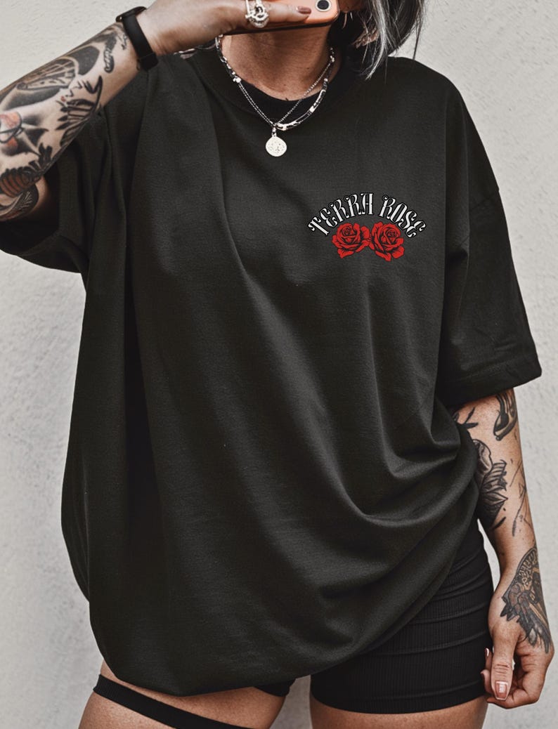 Peut inclure: T-shirt noir surdimensionn&eacute; avec le texte "Terra Rose" au-dessus de deux illustrations de roses rouges. Le t-shirt a des manches courtes et une coupe ample. Le motif est centr&eacute; sur la poitrine.