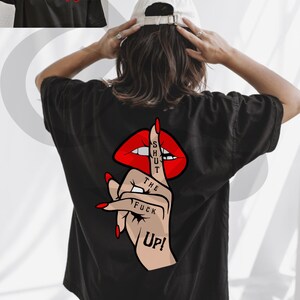 Peut inclure: T-shirt noir avec un motif au dos repr&eacute;sentant des l&egrave;vres rouges, un doigt faisant chut, et les mots "SHUT THE FUCK UP!". Le devant du t-shirt pr&eacute;sente un petit motif de rose rouge avec du texte. Une casquette de baseball blanche est port&eacute;e.