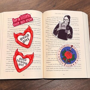 May include: Open book with several stickers. One sticker reads "be a lilith not an eve". Another sticker says "good girl" inside a heart. A third sticker says "good fucking girl" inside a heart. A fourth sticker shows a grim reaper reading a book. A fifth sticker says "enemies to lovers book club".