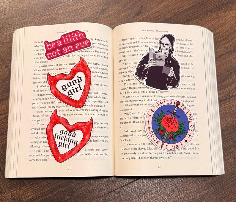 May include: Open book with several stickers. One sticker reads "be a lilith not an eve". Another sticker says "good girl" inside a heart. A third sticker says "good fucking girl" inside a heart. A fourth sticker shows a grim reaper reading a book. A fifth sticker says "enemies to lovers book club".