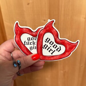 May include: Two heart-shaped stickers with red glitter borders. One sticker reads "good girl" in gothic font, the other reads "god fuckin' girl". The stickers are held in a hand with red nail polish and a silver ring.
